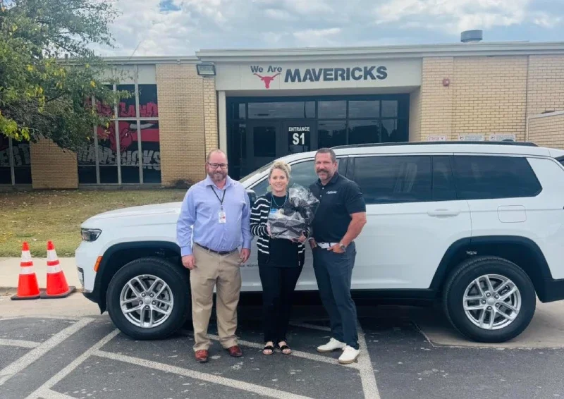 A group of adults stand next to a white SUV in a school parking lot.
