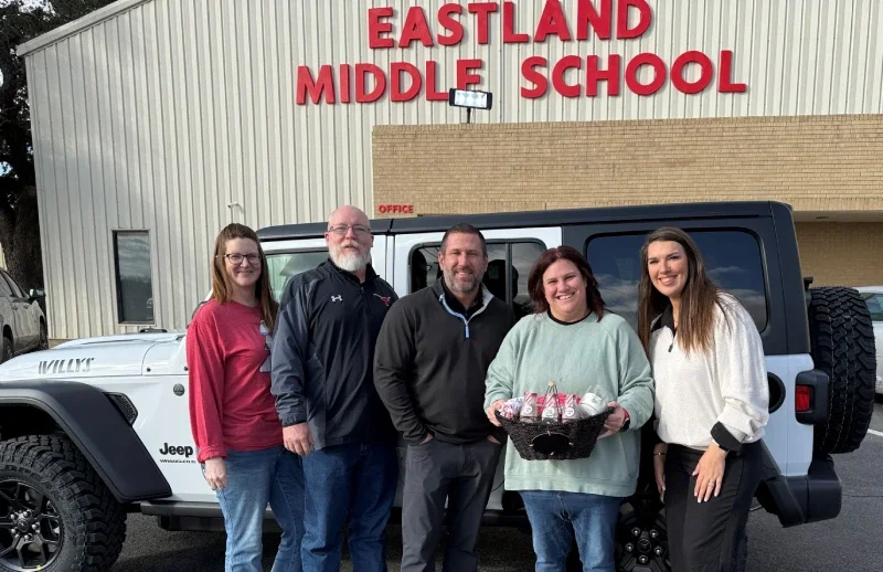 A group of people pose in front of a white Jeep in a school parking lot.